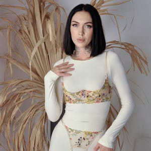 May include: A white long-sleeved top with a floral embroidered bra and matching panty set. The bra and panty feature a delicate floral design in shades of pink, yellow, and blue.