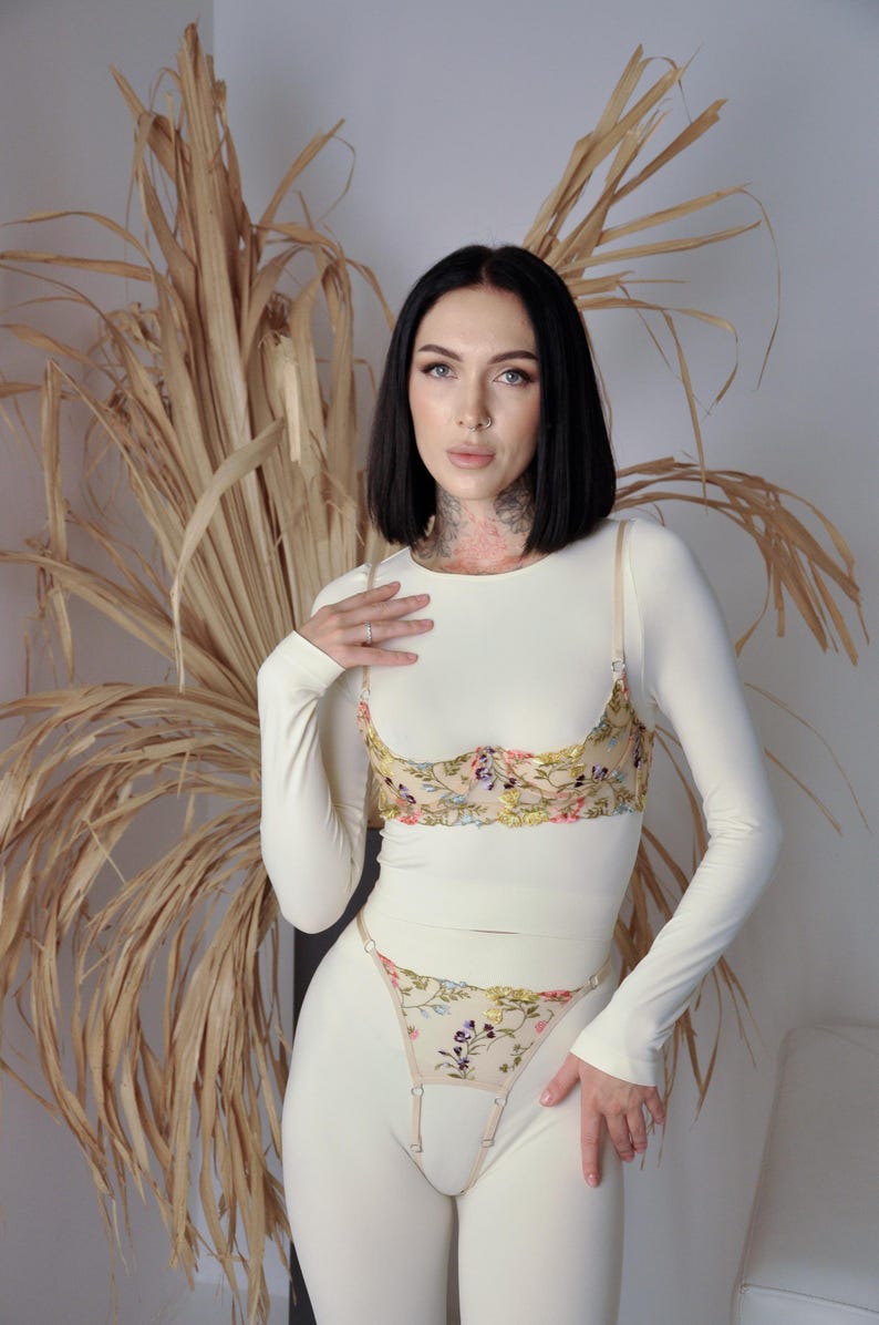 May include: A white long-sleeved top with a floral embroidered bra and matching panty set. The bra and panty feature a delicate floral design in shades of pink, yellow, and blue.