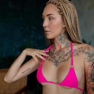 May include: A woman with blonde braids wears a bright pink bikini top with thin straps. She has a floral tattoo on her chest and a tattoo on her arm that says "Forever Young".