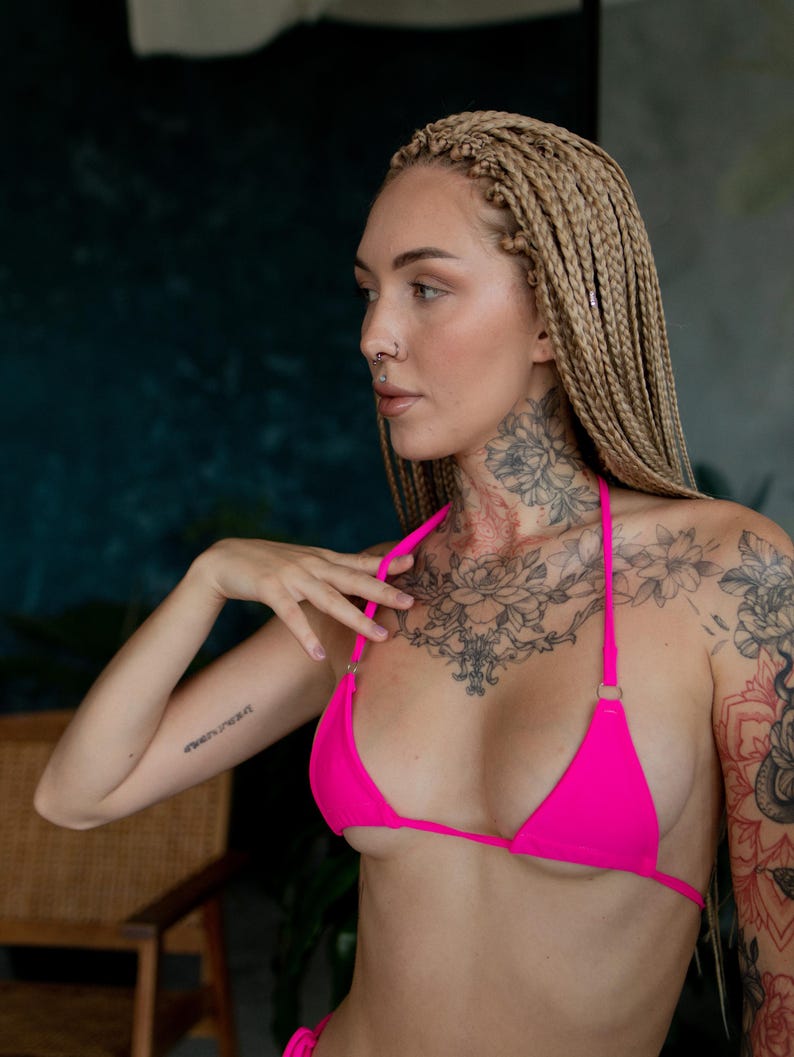 May include: A woman with blonde braids wears a bright pink bikini top with thin straps. She has a floral tattoo on her chest and a tattoo on her arm that says "Forever Young".