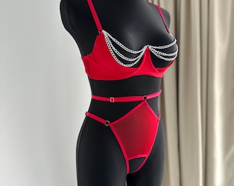 May include: A red lingerie set displayed on a black mannequin. The set includes a bra with silver chain detailing and a matching high-waisted bottom. The bra has adjustable straps. The lingerie is made of a smooth, shiny fabric.