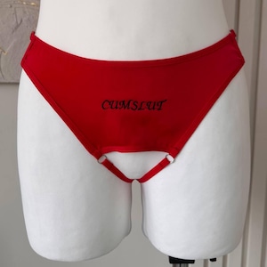 May include: Red underwear featuring the word "CUMSLUT" in black script. The design includes a strap detail and metal rings.