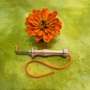 May include: An orange zinnia flower rests above a wooden punch needle tool with a metal needle and orange yarn. The tool has a light wood handle. The yarn is looped in a curved shape. The background is a green, textured fabric.