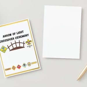 May include: A white card with gold trim and the text "ARROW OF LIGHT CROSSOVER CEREMONY" is displayed next to a blank white card. A gold pen rests on the surface. The card features a bridge graphic and various scout emblems.