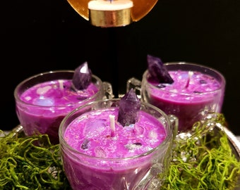 May include: Three purple glass candle holders with amethyst crystals and a purple wax candle inside each. The candle holders are sitting on a silver tray with green moss and purple moss around them.