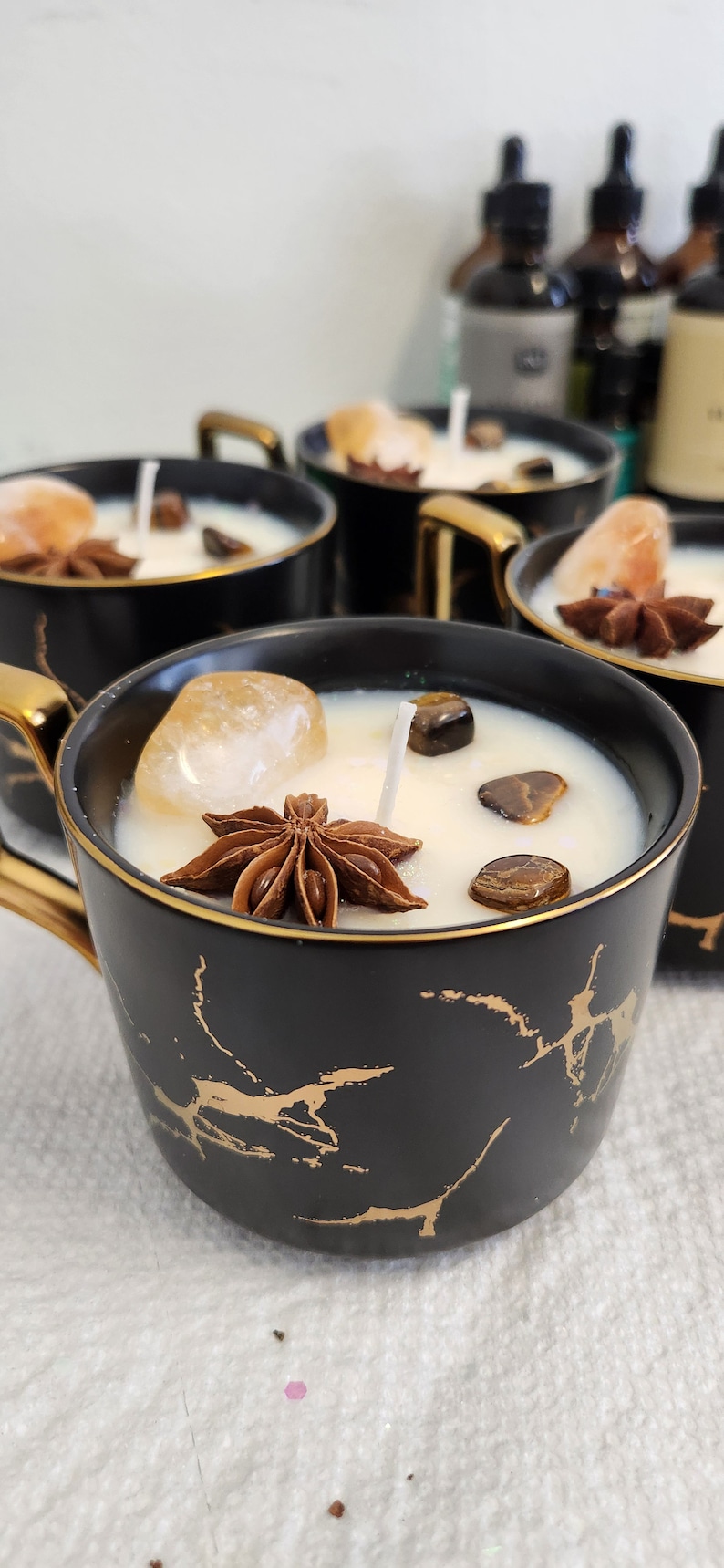 K&ouml;nnte beinhalten: Vier schwarze und goldene Kerzenhalter aus Keramik mit wei&szlig;en Kerzen. Jeder Kerzenhalter hat einen goldenen Rand und einen Sternanis, einen klaren Kristall und Tigeraugen-Steine auf der Kerze.