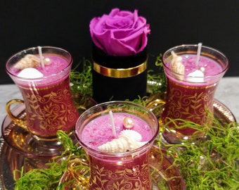 May include: Three decorative candles in ornate glass cups with gold accents, filled with pink wax and topped with seashells and pearls. A vibrant pink rose sits in a black box with gold trim. The candles are on a silver tray with green moss.