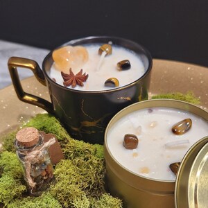 K&ouml;nnte beinhalten: Zwei Kerzen mit eingebetteten Kristallen und eine kleine Flasche mit Kristallen. Die Kerzen befinden sich in einer schwarzen und goldenen Tasse und einer goldenen Dose. Die Kerzen sind von gr&uuml;nem Moos umgeben.