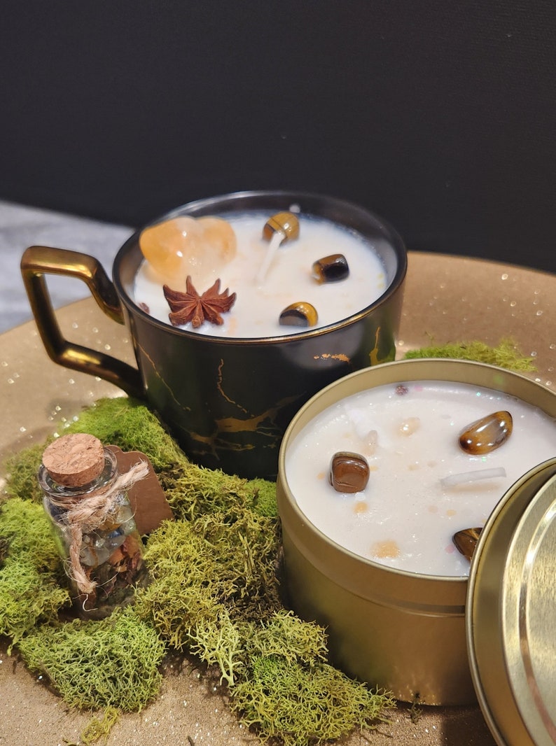 K&ouml;nnte beinhalten: Zwei Kerzen mit eingebetteten Kristallen und eine kleine Flasche mit Kristallen. Die Kerzen befinden sich in einer schwarzen und goldenen Tasse und einer goldenen Dose. Die Kerzen sind von gr&uuml;nem Moos umgeben.