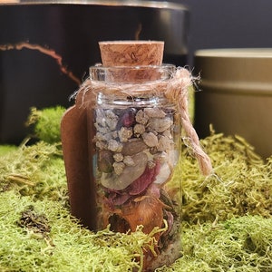 K&ouml;nnte beinhalten: Eine kleine Glasflasche mit einem Korkverschluss, gef&uuml;llt mit verschiedenen Kristallen, Steinen und getrockneten Blumen. Die Flasche ist mit einer Schnur gebunden und hat ein braunes Papieretikett.
