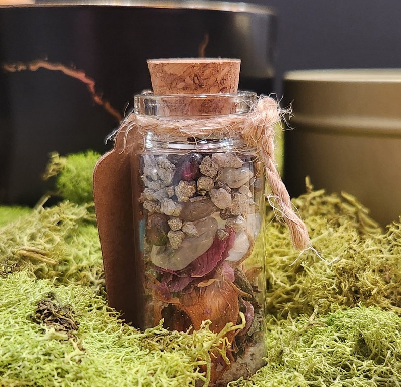 K&ouml;nnte beinhalten: Eine kleine Glasflasche mit einem Korkverschluss, gef&uuml;llt mit verschiedenen Kristallen, Steinen und getrockneten Blumen. Die Flasche ist mit einer Schnur gebunden und hat ein braunes Papieretikett.