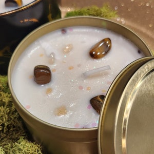 K&ouml;nnte beinhalten: Eine goldene Kerzen-Dose mit Deckel, gef&uuml;llt mit einer wei&szlig;en Kerze und eingebetteten Tigeraugen-Steinen. Die Kerze steht auf einer goldenen Glitzer-Oberfl&auml;che mit gr&uuml;nem Moos.