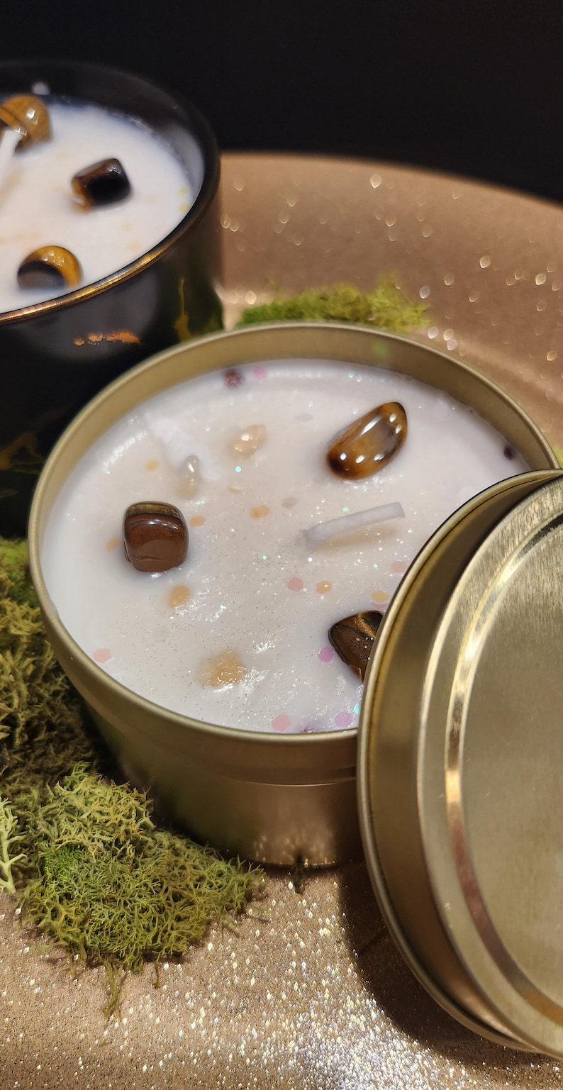 K&ouml;nnte beinhalten: Eine goldene Kerzen-Dose mit Deckel, gef&uuml;llt mit einer wei&szlig;en Kerze und eingebetteten Tigeraugen-Steinen. Die Kerze steht auf einer goldenen Glitzer-Oberfl&auml;che mit gr&uuml;nem Moos.