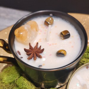 K&ouml;nnte beinhalten: Eine schwarze und goldene Teetassenkerze mit einem wei&szlig;en Wachsoberteil. Die Kerze ist mit einer Sternanis, einem kleinen St&uuml;ck orangefarbenem Calcit und mehreren Tigeraugen-Steinen verziert. Die Kerze steht auf einem Bett aus gr&uuml;nem Moos.