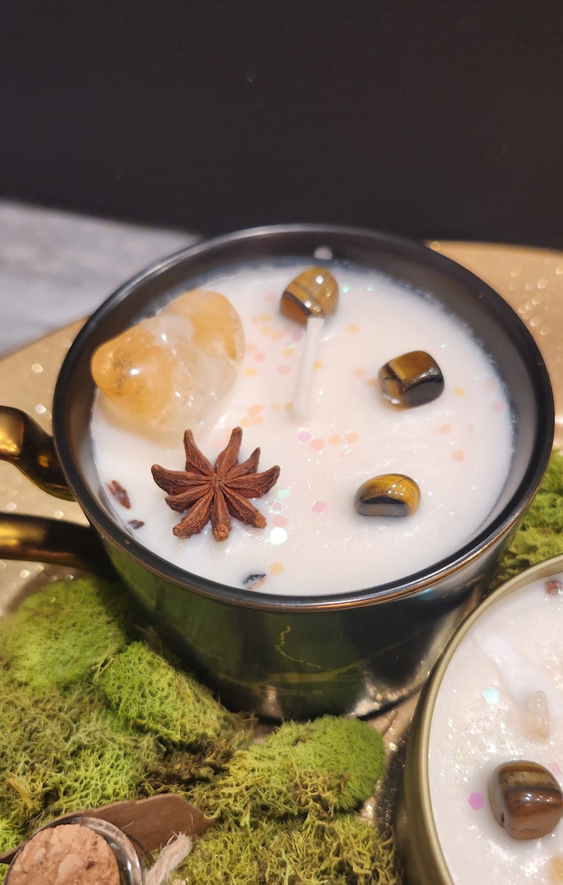 K&ouml;nnte beinhalten: Eine schwarze und goldene Teetassenkerze mit einem wei&szlig;en Wachsoberteil. Die Kerze ist mit einer Sternanis, einem kleinen St&uuml;ck orangefarbenem Calcit und mehreren Tigeraugen-Steinen verziert. Die Kerze steht auf einem Bett aus gr&uuml;nem Moos.