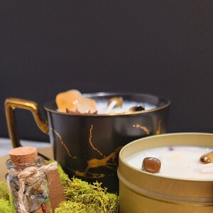 K&ouml;nnte beinhalten: Zwei Kerzen, eine in einer schwarzen und goldenen Tasse und eine in einer goldenen Dose, mit einem kleinen Glasgef&auml;&szlig; gef&uuml;llt mit Kristallen und Moos.