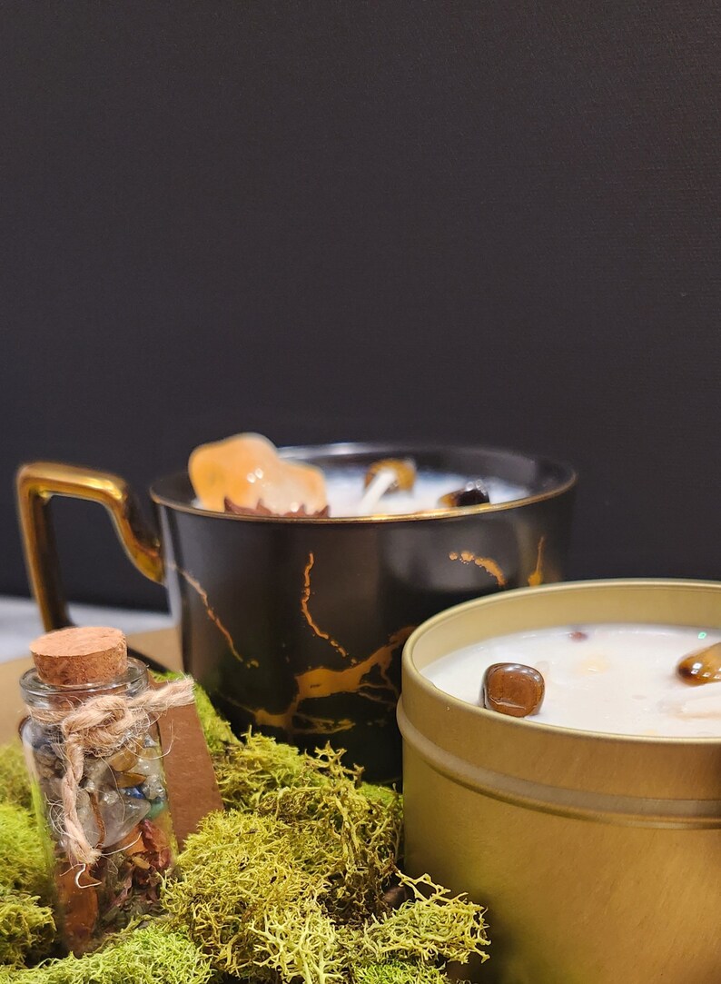 K&ouml;nnte beinhalten: Zwei Kerzen, eine in einer schwarzen und goldenen Tasse und eine in einer goldenen Dose, mit einem kleinen Glasgef&auml;&szlig; gef&uuml;llt mit Kristallen und Moos.