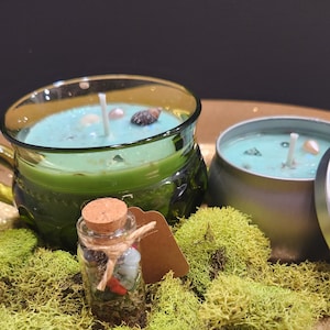 May include: Two teal green glass candle jars with a white wick and a small glass bottle filled with crystals and herbs. The candles are on a gold tray with green moss.