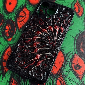 Pode incluir: Capa de telefone preta com um design texturizado e um adorno vermelho e prateado em forma de espinha dorsal. A capa está sobre um fundo verde com ilustrações vermelhas em forma de olho, criando um contraste marcante.