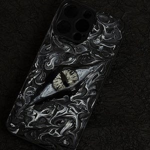 Könnte beinhalten: Schwarz-weiß gemusterte Handyhülle mit detailliertem Augendesign. Die Hülle hat eine glänzende Oberfläche und ein einzigartiges, künstlerisches Muster. Das Auge ist grau und weiß und setzt einen auffälligen Akzent.