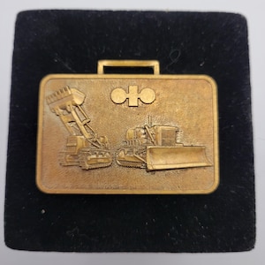May include: A gold-colored metal briefcase-shaped pin featuring embossed images of construction vehicles, including a bulldozer and a front-end loader. The pin has a handle at the top and a logo in the center.