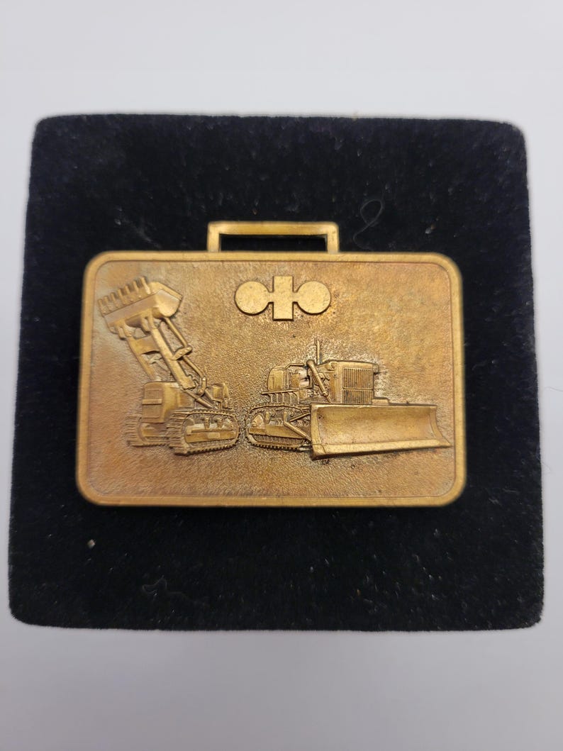 May include: A gold-colored metal briefcase-shaped pin featuring embossed images of construction vehicles, including a bulldozer and a front-end loader. The pin has a handle at the top and a logo in the center.