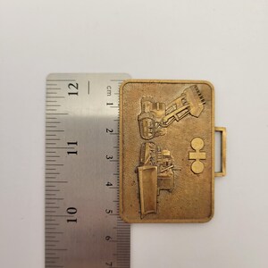 May include: A rectangular, bronze-colored metal badge featuring embossed images of construction vehicles. The badge is approximately 2 inches long and has a small rectangular loop on the right side. The background is a light, neutral color.