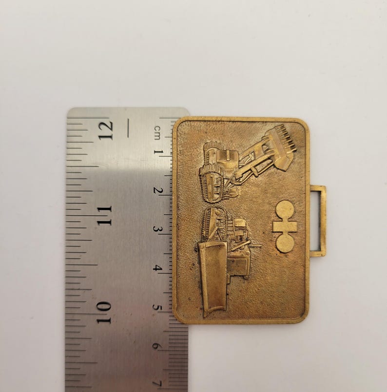 May include: A rectangular, bronze-colored metal badge featuring embossed images of construction vehicles. The badge is approximately 2 inches long and has a small rectangular loop on the right side. The background is a light, neutral color.