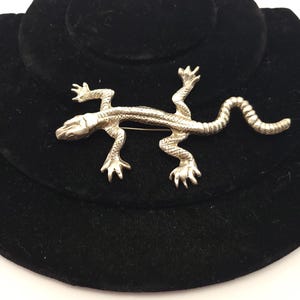 May include: A silver lizard brooch with a detailed design. The lizard is posed in a walking position with its tail curled up.