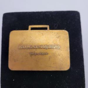 May include: A gold-colored metal briefcase-shaped pin. The pin has the text "KOMATSU, LTD." and "Tokyo, Japan" engraved on it. The pin is set on a black velvet background.