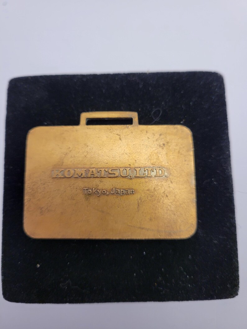 May include: A gold-colored metal briefcase-shaped pin. The pin has the text "KOMATSU, LTD." and "Tokyo, Japan" engraved on it. The pin is set on a black velvet background.