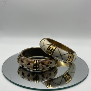 May include: Two decorative bangles, one with a white and gold geometric pattern and the other with a brown and gold floral pattern.