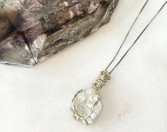 May include: A clear quartz crystal wrapped in gold wire, hanging from a silver chain necklace. The crystal is faceted and has a natural, raw look. The necklace is displayed on a white surface next to a large, clear crystal.