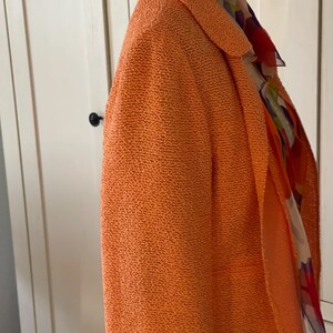 May include: An orange textured blazer with a shawl collar. A colorful patterned scarf is draped over the shoulders. The blazer is displayed on a mannequin. The collar of the blazer is lined with a yellow fabric.