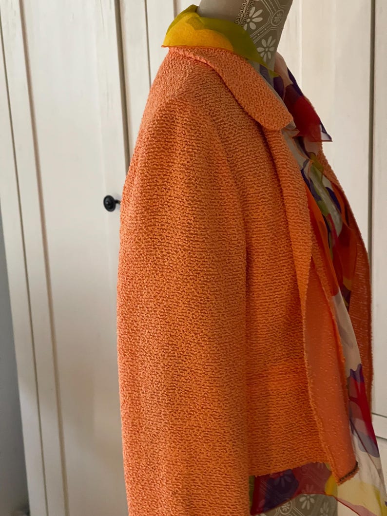 May include: An orange textured blazer with a shawl collar. A colorful patterned scarf is draped over the shoulders. The blazer is displayed on a mannequin. The collar of the blazer is lined with a yellow fabric.
