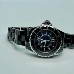 May include: A black ceramic wristwatch with a round face and a black dial. The watch has silver hands and hour markers. The watch band is black ceramic. The watch is displayed on a white surface.