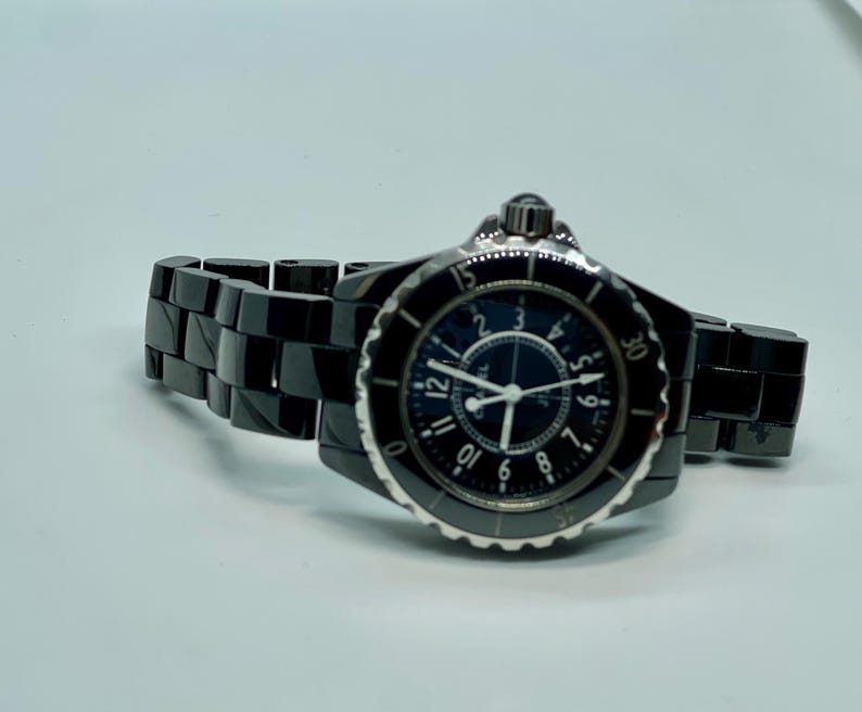 May include: A black ceramic wristwatch with a round face and a black dial. The watch has silver hands and hour markers. The watch band is black ceramic. The watch is displayed on a white surface.