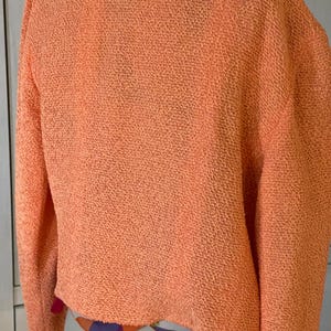 May include: A textured, peach-colored sweater with long sleeves. The sweater has a unique, nubby texture and a slightly cropped fit. The collar and the bottom hem of the sweater reveal a colorful patterned fabric.