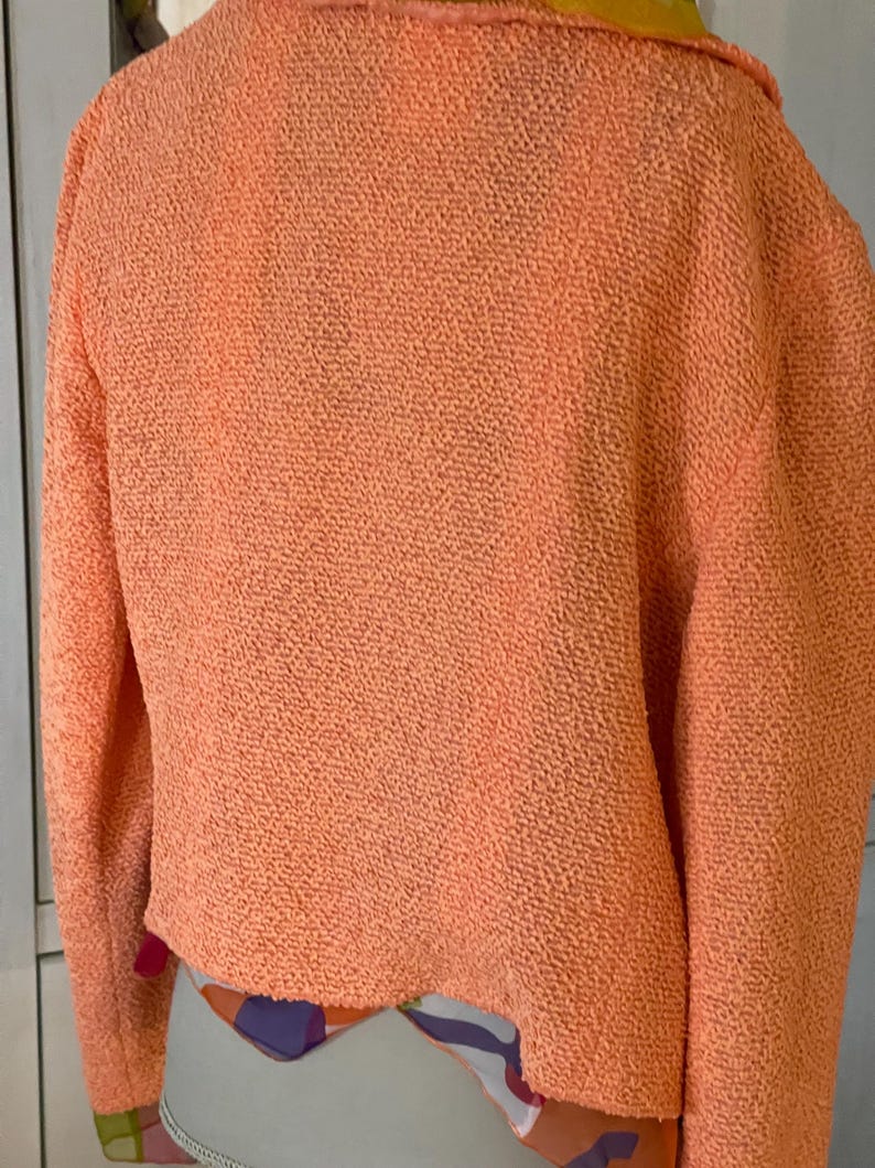 May include: A textured, peach-colored sweater with long sleeves. The sweater has a unique, nubby texture and a slightly cropped fit. The collar and the bottom hem of the sweater reveal a colorful patterned fabric.