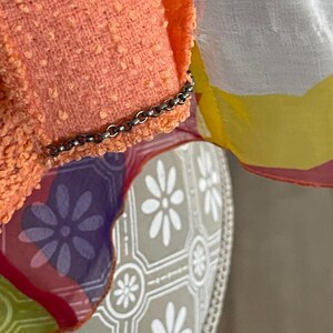 May include: Close-up of a peach-colored textured fabric with a silver chain detail. A sheer fabric with floral patterns in purple, yellow, and white is visible. The background features a patterned surface with white flower designs.