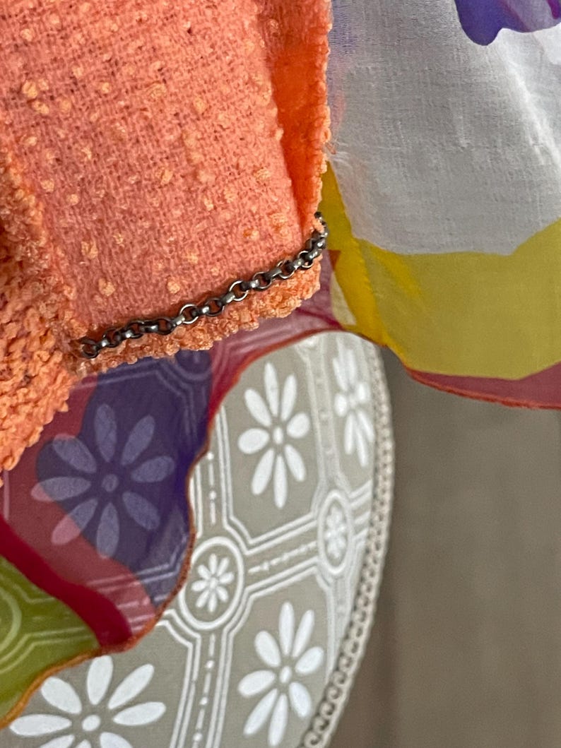 May include: Close-up of a peach-colored textured fabric with a silver chain detail. A sheer fabric with floral patterns in purple, yellow, and white is visible. The background features a patterned surface with white flower designs.