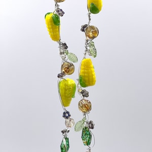 May include: A handmade necklace featuring yellow corn, brown and clear beads, green leaves, and silver wire. The necklace has a whimsical design with a farm-to-table theme. The corn is a bright yellow with green accents.