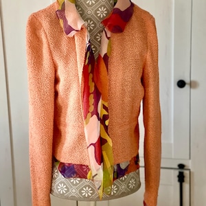 May include: A peach-colored textured jacket with long sleeves and a colorful floral scarf. The scarf features a mix of red, purple, and yellow patterns. The jacket is displayed on a mannequin.