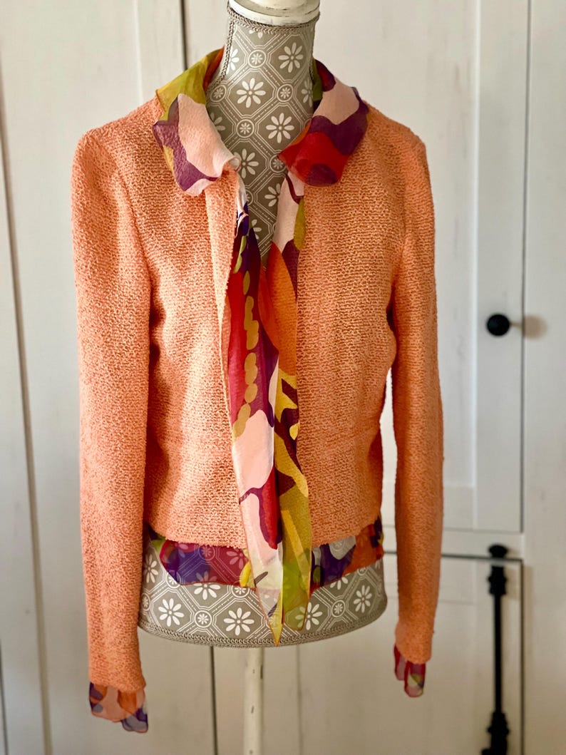 May include: A peach-colored textured jacket with long sleeves and a colorful floral scarf. The scarf features a mix of red, purple, and yellow patterns. The jacket is displayed on a mannequin.