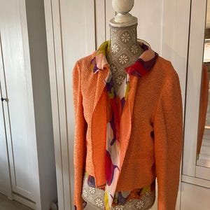 May include: An orange textured jacket with long sleeves and an open front is displayed on a mannequin. A colorful scarf with floral and abstract patterns is draped around the neck. The mannequin has a decorative floral pattern.