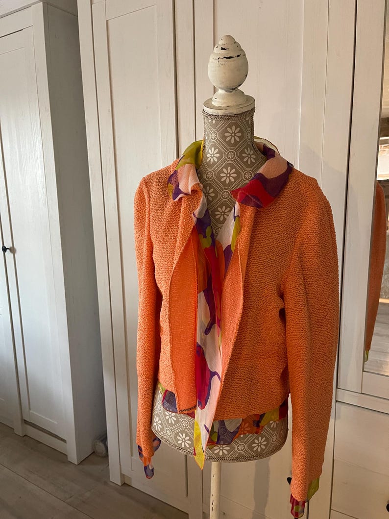 May include: An orange textured jacket with long sleeves and an open front is displayed on a mannequin. A colorful scarf with floral and abstract patterns is draped around the neck. The mannequin has a decorative floral pattern.