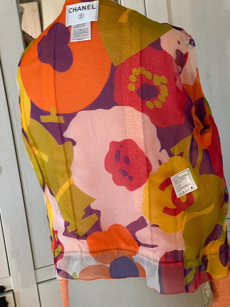 May include: A sheer, floral-print top with a vibrant mix of colors including orange, pink, yellow, and purple. The top features a large floral pattern and a tag that reads "CHANEL". The garment is lightweight and appears to be made of a delicate fabric.