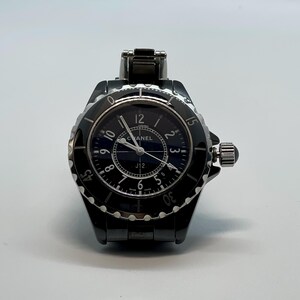 May include: A black Chanel J12 watch with a round face and a black bezel. The watch has a dark blue dial with white numerals and hands. The watch band is black and made of a link style material. The watch is on a white background.