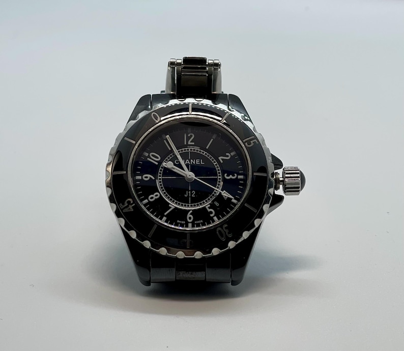 May include: A black Chanel J12 watch with a round face and a black bezel. The watch has a dark blue dial with white numerals and hands. The watch band is black and made of a link style material. The watch is on a white background.