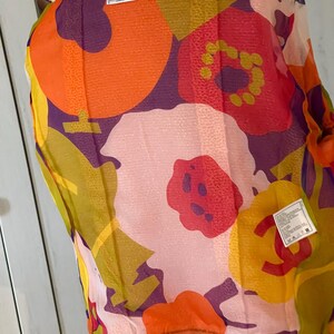 May include: A sheer scarf with a vibrant floral pattern in shades of orange, red, purple, yellow, and green. The scarf has a white border and a tag with the brand name "CHANEL".